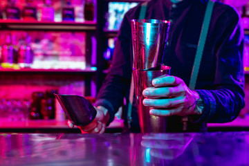 Close up of professional barman finishes preparation of alcoholic cocktail in multicolored neon light, gives it to client. Entertainment, drinks, service concept. Modern bar, trendy neoned colors.