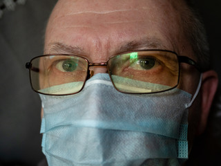 Close-up face portrait of senior man in medical mask and glasses looking at camera indoors