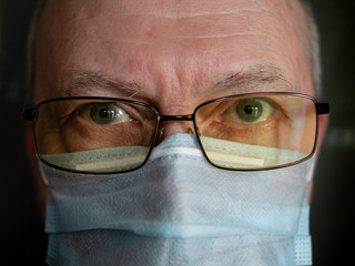 Close-up face portrait of senior man in medical mask and glasses looking at camera indoors
