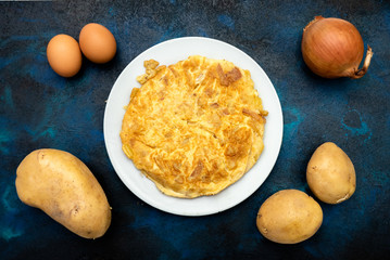 potato omelette on white plate with ingredients. Eggs, potato and onion, on blue and black background