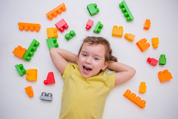 A beautiful little girl lies on a white background with a constructor and smiles. A cute girl in a yellow t-shirt with ponytails laughs while looking at the camera. Children's game. Training.