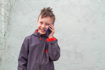 The boy stands against the wall of the house and happily talks on a blue mobile phone. Fall, spring, or cold summer.