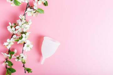 Reusable white eco menstrual cup on a pink background, spring cherry tree branches with white flowers. Ecology and recycling concept, zero waste. Women's hygiene, menstruation, critical days.