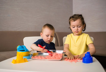 A little girl and a boy sit at a table and play with kinetic sand with a rake, a shovel and different forms of sand. Games at home for children. Games for children's sensory development.