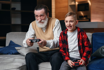 Cute little boy with grandfather sitting on sofa and playing video game with game pad