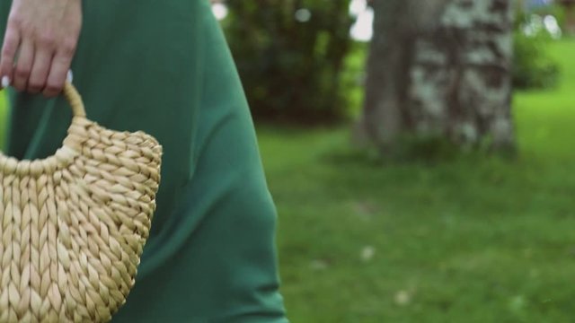 Close up of young woman with handmade stylish straw handbag walks in the summer park. Eco bag.