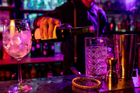 Close up of professional barman finishes preparation of alcoholic cocktail in multicolored neon light, gives it to client. Entertainment, drinks, service concept. Modern bar, trendy neoned colors.
