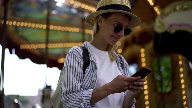 Smiling Young Woman Tourist In Black Sunglasses Chatting Online In Social Networks On Smartphone Device Using Public 4G Internet Connection Standing Outdoors With Carousel On Background
