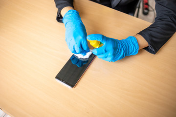 Woman's hand in blue gloves sanitizing cleaning smartphone mobile phone on wood table surface with wet wipes