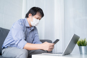 Social distancing self isolation quarantine lockdown, Asian businessman work from home during coronavirus covid-19 disease,wearing protective facemask, living room using phone laptop computer working