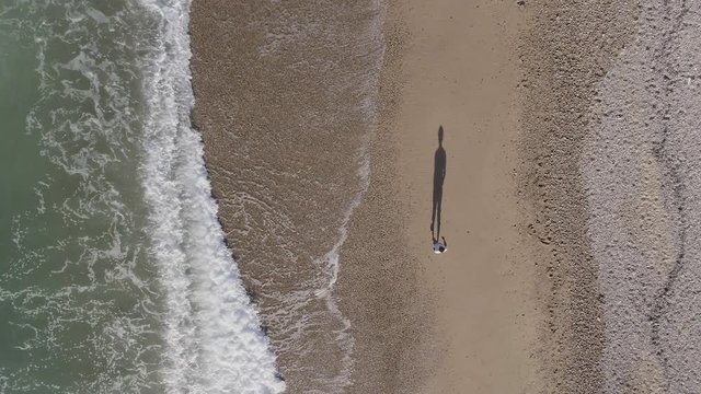 AERIAL DRONE TOP DOWN ZENITHAL SHOT OF MAN WALKING ALONE IN THE SHORE OF THE BEACH WITH BIG SHADOW AND STRONG TIDE