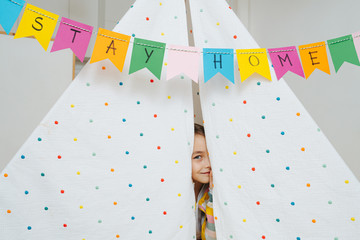 Cute little girl peeking out from a hut with stay at home flag garland across