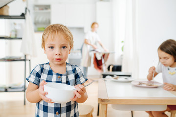 Portrait of toddler boy standing with bowl in hands, sister eating, mom ironing