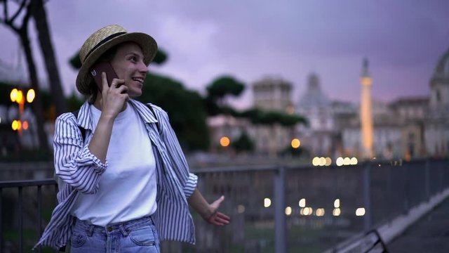 Slow motion effect, smiling hipster girl walks down city street and happily talks on mobile phone, while sharing her travel impressions. Cheerful Caucasian female calling via smartphone outdoors 
