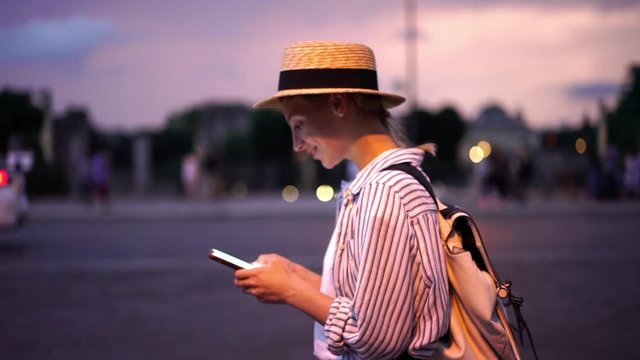 Slow motion of cheerful Caucasian hipster girl typing text message on smartphone device while walking in evening city during travel. Smiling young millennial female chatting online on cellphone

