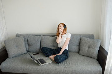 Fototapeta na wymiar Redhead girl sitting on a sofa in home clothes and headphones with laptop and yawns widely, hiding behind hand, frame from above