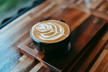 Black cup of hot cappuccino with beautiful latte art on wooden table.