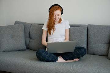 Redhead girl sitting on a sofa in home clothes with a laptop on crossed legs and smiling, looking down © bestsenny