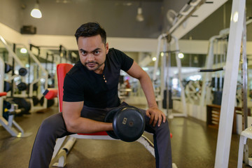 Full body shot of young bearded Indian man lifting weights at the gym