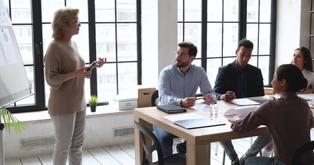 Happy skilled middle aged female business trainer educating motivated young diverse employees, answering on questions at workshop. Smiling businesswoman presenting project details to coworkers.