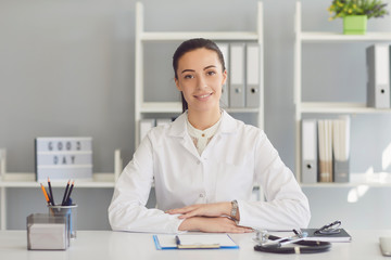 Positive doctor woman looking at the camera smiling while sitting at a table in a clinic.