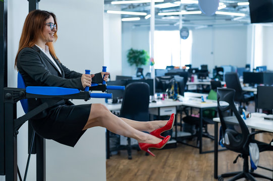 Happy red-haired business woman doing press exercises on a mounted horizontal bar in the office. A female employee in an official suit does sports at the workplace during the break.