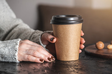 Women's hands in a warm sweater are holding a paper cup of aromatic coffee. The nails are coated with brown nail polish. Coffee manicure. Breakfast or a break in a cozy cafe