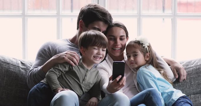 Emotional mixed race mother cuddling adorable small kids with husband, posing for family photo on smartphone indoors. Happy parents recording funny video on cellphone with little children siblings.