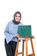 Beautiful hijab woman standing on the side of the chair while holding a shopping bag with expressions of, smiling, isolated on white background