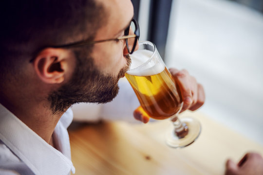 Closeup of handsome unshaven man in shirt sitting in pub next to window, drinking beer and relaxing after work.