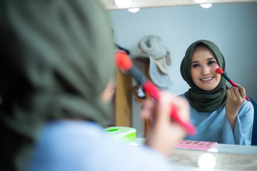 Beautiful hijab woman is dressing up in front of the mirror while holding brush. Hijab girl at makeup room.