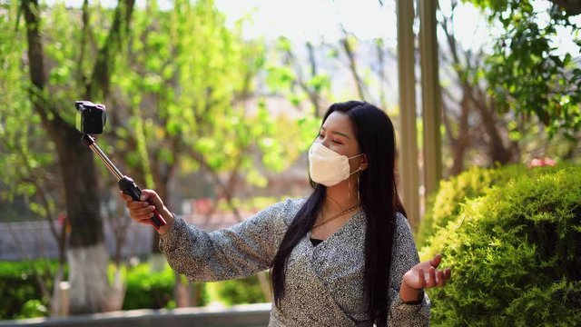 Chinese girl vlogs outside with camera and face mask. China woman with selfie stick records video blog about virus walking outside in park for viewers wearing mask.