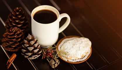Sweet sugar powder cookies, pine cones and a white mug with black coffe on the background of a wood table. Cozy holidays.