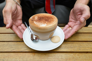 Businessman holding cup of cappuccino coffee with chocolate sprinkles on foam and biscuit on the side