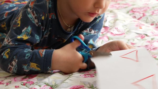 A little boy carefully cuts paper geometric figures with scissors lying down on the bed