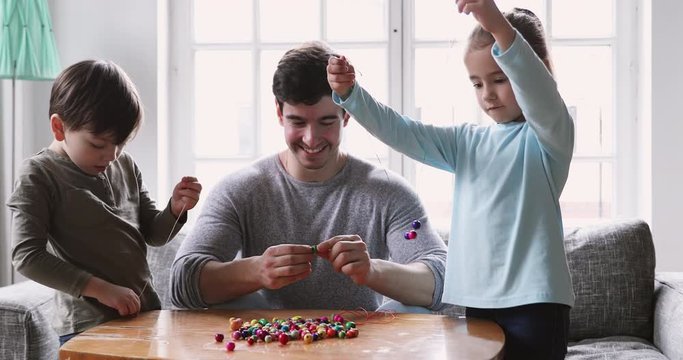 Happy young dad enjoying hobby time with kids siblings, creating handmade accessories with wooden bead pieces. Smiling adorable children involved in creative activity with loving father at home.