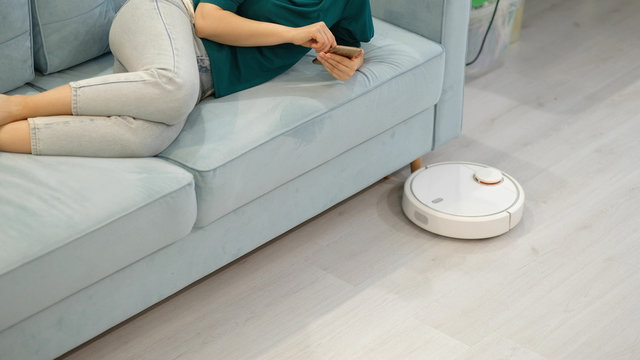 Robotic vacuum cleaner cleaning the room while woman resting on sofa.