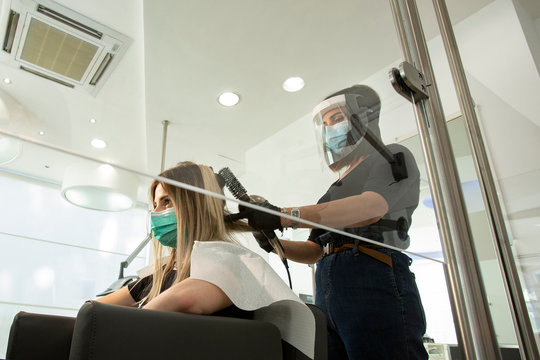 parrucchiera si accinge ad asciugare con un phon i capelli di una di una cliente all'interno di un salone. Entrambi sono equipaggiati di protezioni per evitare contatti e contaminazioni reciproche