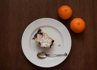 A bitten piece of grated shortcake with jam and a silver spoon on a white plate and two tangerines on a wooden table. Top view