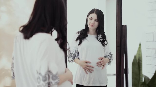 A pregnant young attractive girl, brunette, with long straight hair and a white shirt, looks at herself and admires her belly in the mirror.