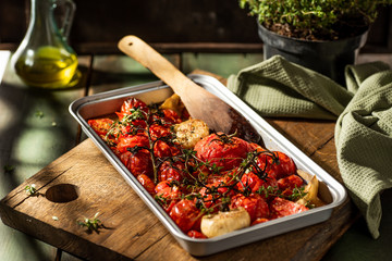 Tomatoes roasted on baking tray with garlic head and thyme. Rustic background