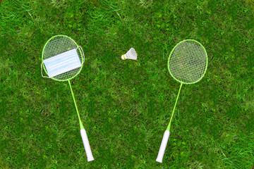 Coronavirus protection symbol. Badminton rackets and shuttlecock on green grass. Medical mask on one racket. view from above