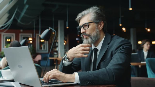 Serious businessman in formal clothing and glasses is working with laptop in cafe then touching beard thinking sitting at table alone. People and business concept.