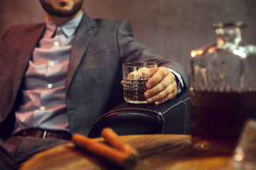 Elegant man with the glass of whisky and two cigars and a carafe of whisky on wooden table in the...