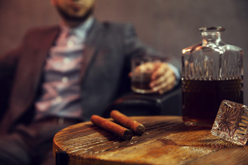 Two cigars and a carafe of whisky on wooden table and an elegant man with the glass of whisky in...