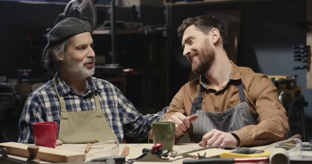 Portrait shot of two cheerful Caucasian craftsmen in goggles, older and younger, smiling in workshop. Senior happy father with handsome son in glasses giving thumbs up to camera in workroom.