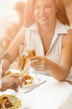 Happy woman smiles holding glass of white wine. Mature couple have event outdoors at beach cafe. Selective focus on human hands with glasses of wine or champagne in foreground. Toned image