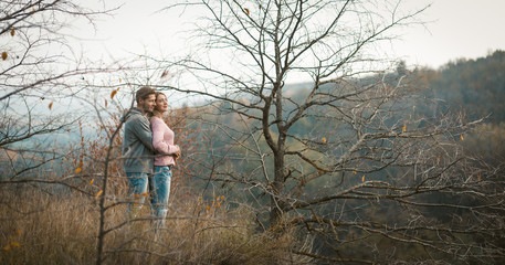 Fototapeta premium Hugging couple in love stands on a hillside looking down, young man and a woman admire the autumn forest growing on the slopes below