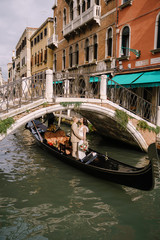 Italy wedding in Venice. A gondolier rolls a bride and groom in a classic wooden gondola along a narrow Venetian canal. Newlyweds in a gondola swim out from under the bridge, stand in a boat and hug.