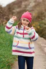 Happy young girl in colorful coat, pink hat, black trousers jumping and running on seaside